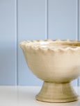 Cream ceramic pedestal urn with scalloped edge and crackle glaze finish, styled against pale blue wall panelling in a Hamptons-style interior.