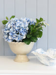 Cream ceramic pedestal urn filled with blue hydrangeas styled beside a blue and white patterned decorative bowl on a shelf in a Hamptons-inspired home.