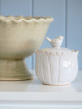 White ceramic lidded jar with bird finial detail, styled on a shelf with a cream pedestal urn for a soft coastal Hamptons look.