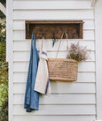 Wooden wall shelf with hooks styled on white weatherboard wall, holding linen, denim and a woven basket
