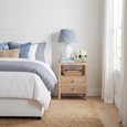 Arden Whitewash Oak Side Table styled beside a white upholstered bed with blue and white bedding, a table lamp and white hydrangeas in a coastal Hamptons bedroom.