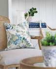 Blue Sapphire floral cushion styled on a classic cane-back armchair with a soft upholstered seat, set against a bright white coastal bookshelf vignette.