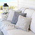 Neutral coastal outdoor sofa styled with striped cushions, a coral motif cushion and soft fringed throw beside a lantern on a Hamptons style verandah