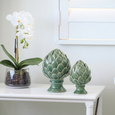 Large and small green Cynara artichoke sculptures styled on a white coastal console table beside a white orchid