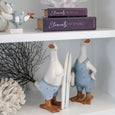Decorative surfing duck figures styled on a white bookshelf with coral sculptures and coastal decor, creating a playful Hamptons-inspired display.