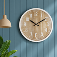 East Hamptons Wall Clock 60cm with a weathered natural timber-look face, white numerals and black hands, styled on a blue panelled wall.