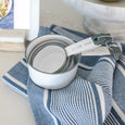 Blue striped Ellis tea towel styled with white measuring cups on a kitchen bench beside marble decor.