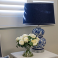 Hamptons style bedside table with blue and white ceramic lamp, navy shade and white roses in glass vase, styled in a calm coastal bedroom setting with plantation shutters