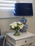 Hamptons style bedside table with blue and white ceramic lamp, navy shade and white roses in glass vase, styled in a calm coastal bedroom setting with plantation shutters