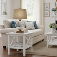 Harbour Side Table White styled beside a white sofa in a bright coastal living room, featuring cross-detail sides, a lower shelf, and a glass table lamp.
