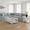 Harbour Side Table White styled beside a pale blue sectional sofa in a bright coastal living room, featuring white cross-detail sides, a lower display shelf, and a ceramic table lamp.