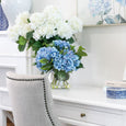 White hydrangea bundle styled in a glass vase on a white console table beside blue hydrangeas in a calm Hamptons-inspired interior.