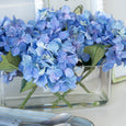 Blue faux hydrangea arrangement in a glass trough vase styled with coastal decor on a white console.