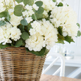 Lifelike white hydrangea mixed bouquet styled in a woven rattan basket on a timber dining table in a relaxed Hamptons-inspired interior.