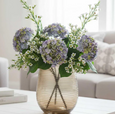Blue hydrangea stems arranged in a glass vase with delicate white florals on a coffee table in a light-filled living room.