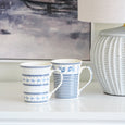 Two Le Petite blue and white ceramic mugs with coordinating striped and floral patterns styled on a Hamptons console beside a textured ceramic lamp and coastal artwork.