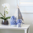 LED sailboat with striped navy and white sails styled on a white console table beside blue and white owl ornaments and a white orchid, creating a coastal Hamptons-inspired display.