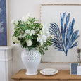 White lilac faux floral arrangement styled in a white ginger jar on a coastal console table with blue botanical artwork.