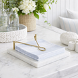 A marble napkin holder with a brass arm holding soft blue napkins on a white marble surface with woven and coastal decor