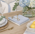 A marble and brass napkin holder on a coastal dining table with blue striped linen napkins, hydrangeas, and lemons