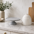 Grey marble footed cake stand styled on a marble kitchen benchtop beside ceramic canister and timber serving board in coastal kitchen