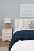 Natural rattan table lamp with sculptural woven base and white shade on bedside table in relaxed coastal bedroom
