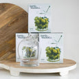 Glass olive jar with spoon styled on a serving board alongside entertaining accessories and gift packaging in a kitchen setting