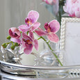 Purple faux orchid stem in a clear glass vase styled on a silver tray beside decorative accessories