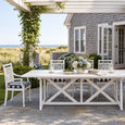 Portofino White Outdoor Dining Table styled with coordinating white cross-back chairs, navy striped cushions and hydrangeas on a coastal patio overlooking the sea.