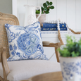 Portsea blue and white floral cushion styled on a classic cane-back armchair with a soft upholstered seat, set against a bright white coastal bookshelf vignette.