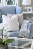 Soft coastal armchair styled in light blue upholstery, layered with neutral cushions and a pale blue shell-pattern throw draped naturally over the arm, set beside a white side table with coastal décor and a glass coffee table with white orchid in the foreground.