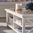 Styled close-up of the Sorrento Hamptons white coffee table featuring glass top, shelf storage, books, woven basket and coastal decor accents