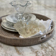 Verandah Round Rattan Tray styled on a marble benchtop with layered white ceramics, decorative white coral and folded linen napkins.