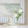 White lisianthus stems arranged in a clear glass bottle vase beside a butterfly ceramic vase and coastal artwork in a soft Hamptons-style interior.