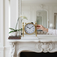 White Marble & Gold Column Mantel Clock styled on an ornate white fireplace mantel beside a potted orchid and decorative books in an elegant classic interior.