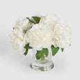 White faux peony arrangement with full layered blooms and green leaves presented in a clear glass water-effect vase.