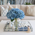 White wooden hanging beads styled on a mirrored tray beside blue hydrangeas in a coastal living room.